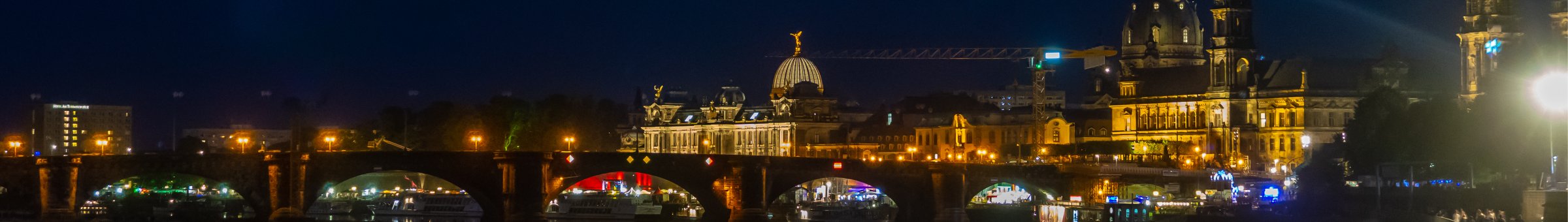 Ein Foto ist dargestellt vom Ufer der Elbe aus, auf die beleuchtete Landeshauptstadt Dresden Altstadt und die Albertbrücke. Angestrahlt glänzt die Stadt bei Nacht in besonders goldenem Licht.