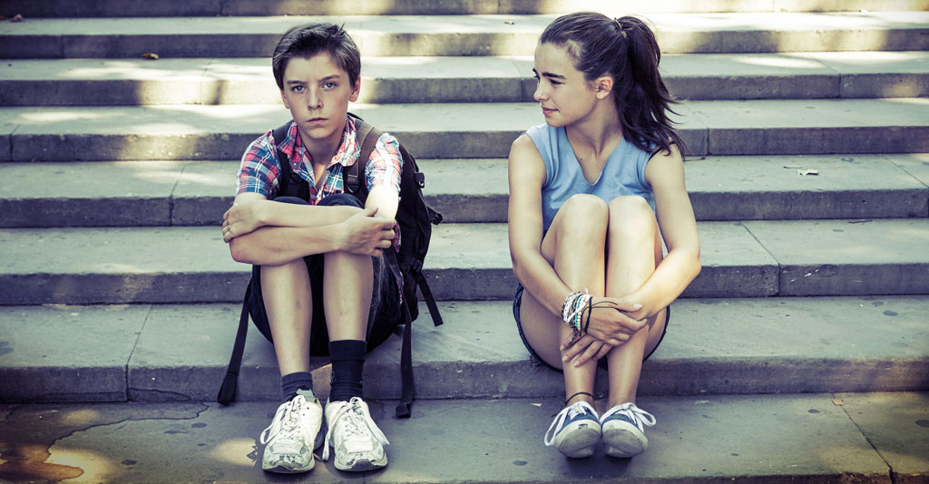 Zwei Jugendliche, ein Junge und ein Mädchen sitzen auf einer Treppe. Beide umschlingen in der Sitzposition jeweils die eigenen Beine. Der Junge schaut emotionslos geradeaus, während das Mädchen seinen Blick aufmerksam in seine Richtung lenkt.