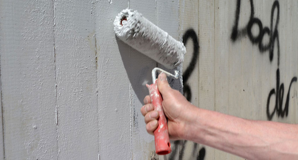 Jemand übermalt eine Schmiererei an der Wand mit einer Malerrolle und grau-weißer Farbe.